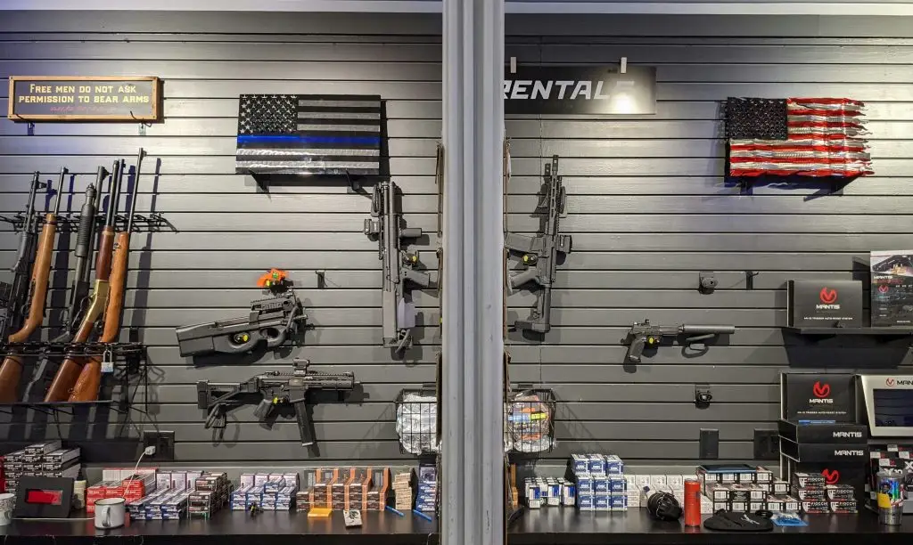 Indoor range rental wall displaying rifles and pistols on gray slat wall; ammo and Mantis training boxes below; thin blue line and distressed U.S. flag art above.